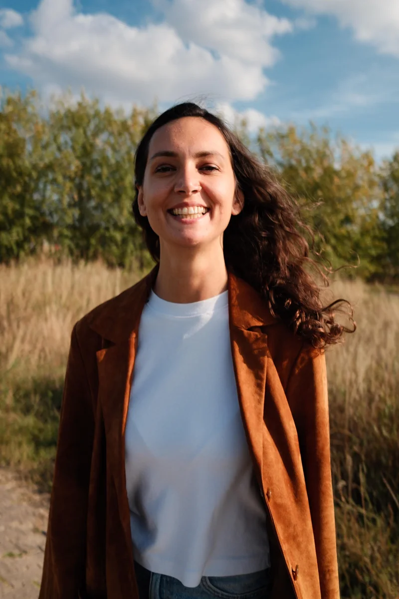 Benan facing the camera outdoors, wearing a brown jacket, white T-shirt and jeans