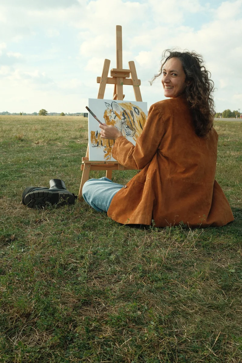 Benan seated beside her easel, holding a brush and looking back toward the camera